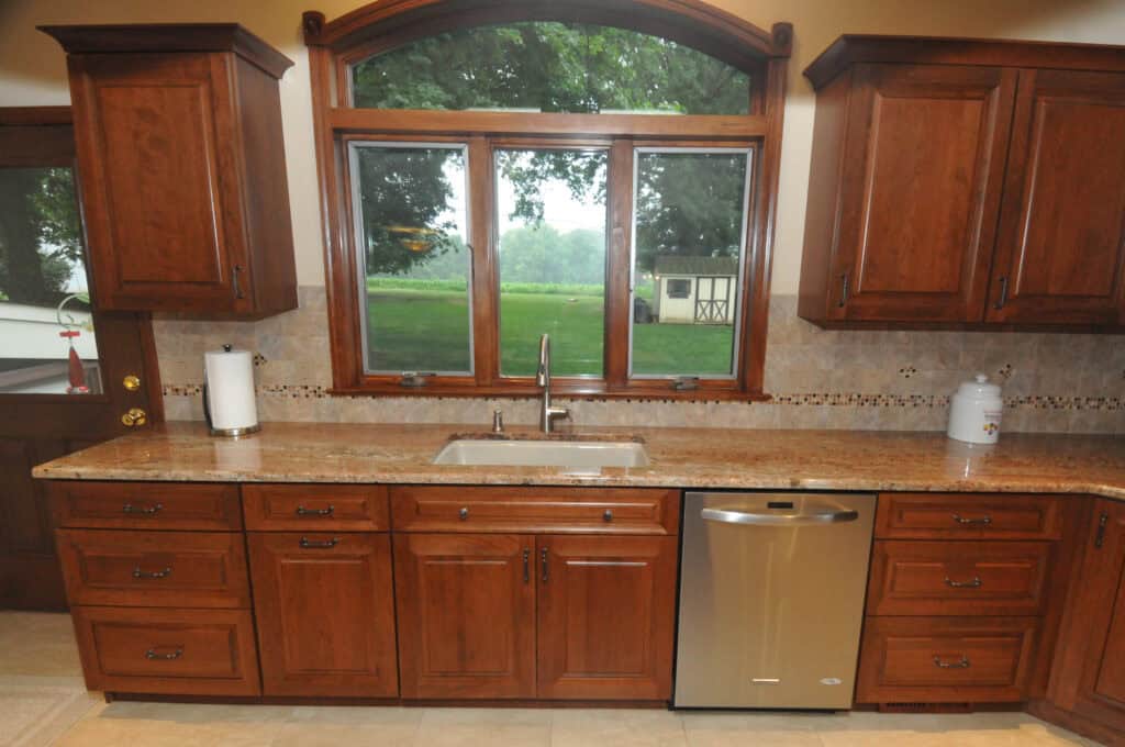 A kitchen with brown wooden cabinets, a granite countertop, a sink under a large window, a stainless steel dishwasher, and a paper towel holder on the counter; green trees and grass visible outside.
