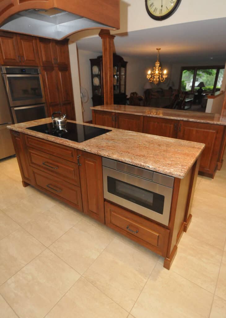 A modern kitchen with wooden cabinets, granite countertop island featuring a stovetop and built-in microwave, tiled floor, and dining area with chandelier in the background.