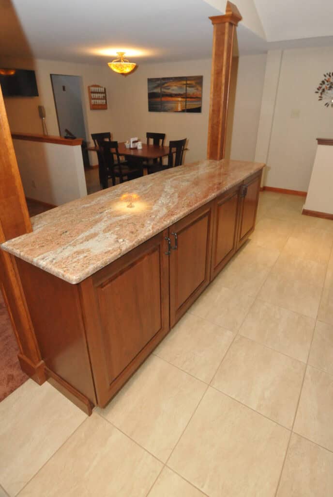 A kitchen island with a polished granite countertop and wooden cabinets stands on a tiled floor, with a dining table and chairs visible in the background.