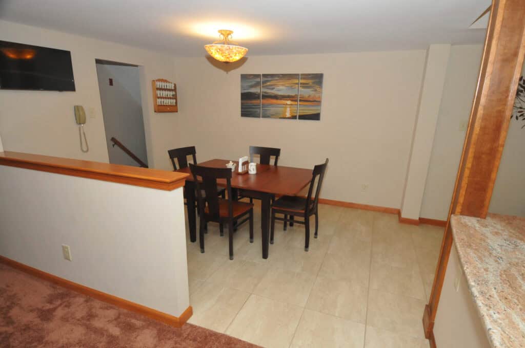 A dining area with a square wooden table and five chairs, a wall-mounted phone, a large wall art of a sunset, tile flooring, beige walls, and a decorative light fixture overhead.