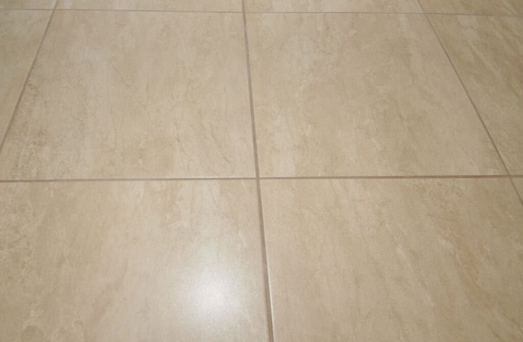 Beige ceramic floor tiles with subtle marble-like veining, arranged in a square grid pattern with thin grout lines. The surface has a slight sheen, reflecting light.