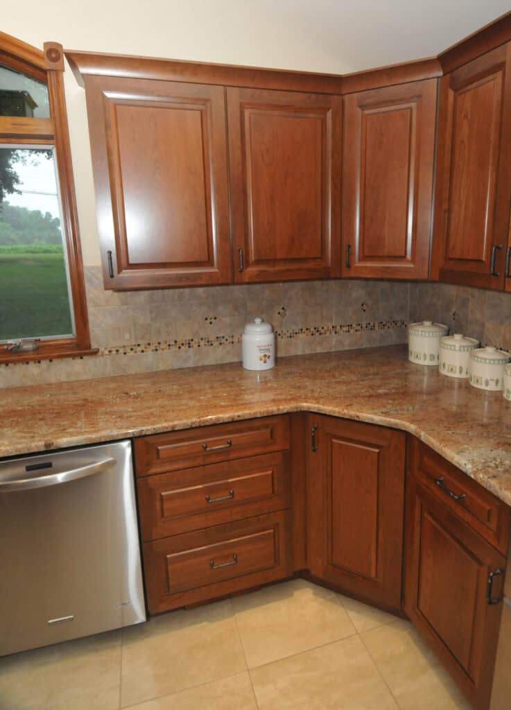 A kitchen corner with brown wooden cabinets, granite countertops, beige tile backsplash with a decorative strip, a stainless steel dishwasher, and ceramic canisters on the counter near a window.