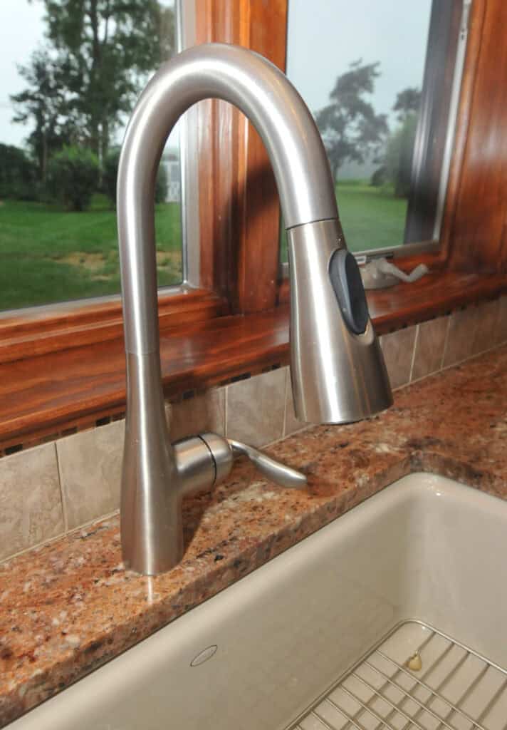 A modern stainless steel kitchen faucet with a curved neck and pull-down spray head is installed on a brown granite countertop next to a white sink, with windows and a green yard in the background.