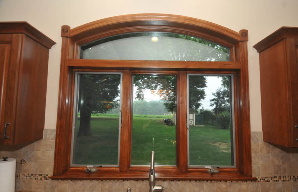 A large kitchen window with a wooden frame overlooks a green lawn and trees. Brown cabinets and beige tiled backsplash are visible on either side, with a silver faucet beneath the window.