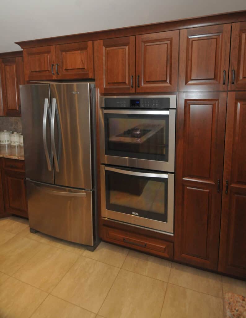 A modern kitchen with wooden cabinets, a stainless steel French door refrigerator on the left, and a built-in double oven in the center, all set against a tiled floor.