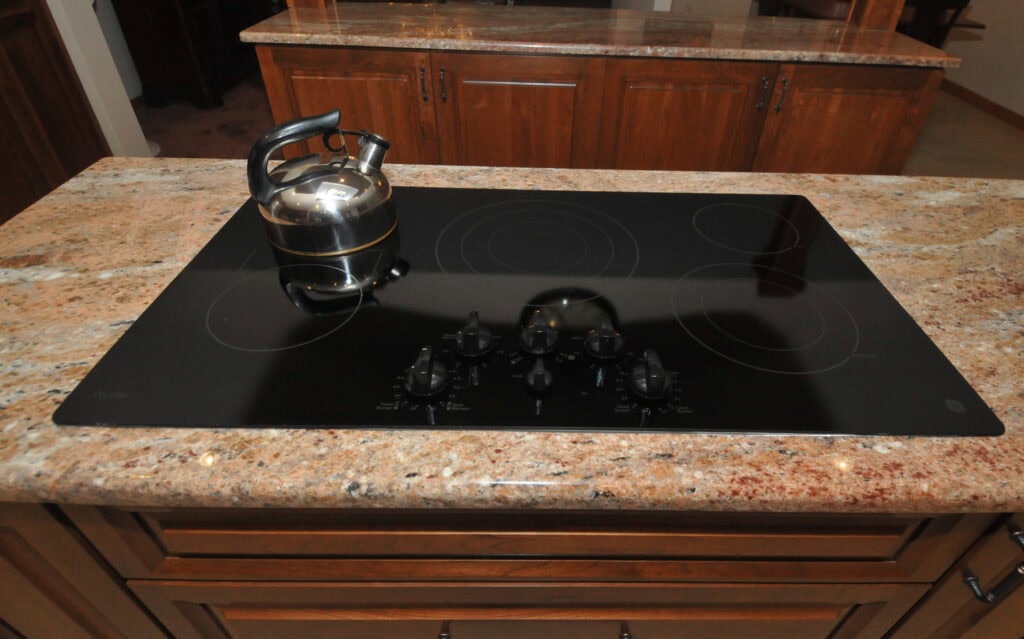 A stainless steel tea kettle sits on a black glass electric cooktop with multiple burner controls, built into a granite countertop in a kitchen with wooden cabinets.