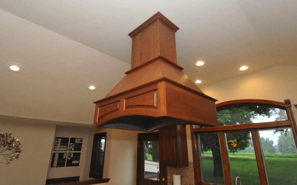 A large, wooden kitchen range hood hangs from the ceiling above a cooking area, with recessed lights and large windows offering a view of greenery outside.