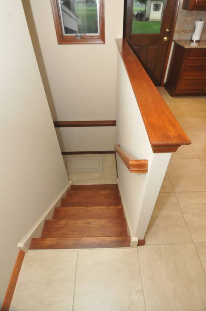 View from the top of a wooden staircase leading down to a small landing, with beige walls, a wooden handrail, and a tiled floor in a kitchen area. A window and wooden door are visible in the background.