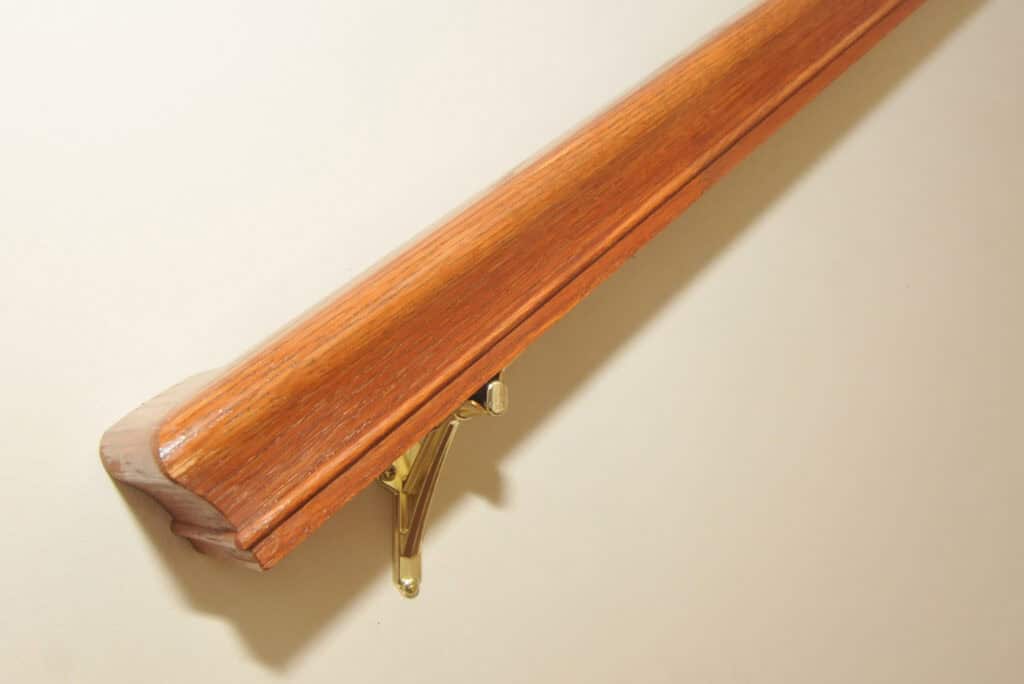 Close-up of a wooden handrail mounted on a beige wall with a brass bracket. The handrail has a polished finish and a beveled edge at the end.