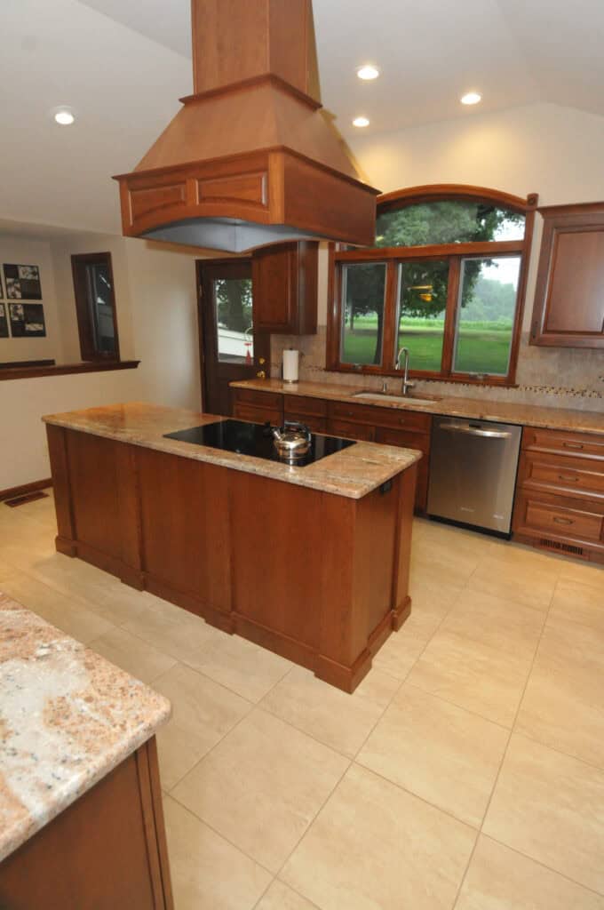 Modern kitchen with wood cabinetry, central island with cooktop, stainless steel dishwasher, granite countertops, tile floor, large window with outdoor view, and recessed lighting in the ceiling.