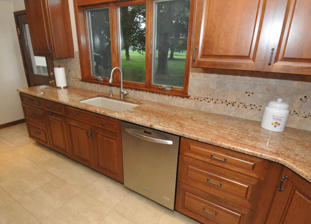 Spacious kitchen with wood cabinets, granite countertops, a stainless steel dishwasher, built-in sink, paper towel holder, and large window overlooking a green yard. A ceramic jar sits on the counter.