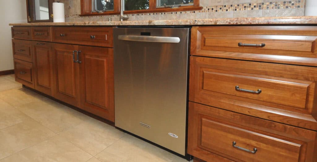 A modern kitchen features wooden cabinets and drawers with dark handles, a built-in stainless steel dishwasher, beige tile flooring, and a stone countertop with a backsplash near a window.