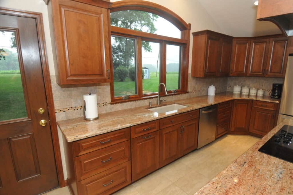 A kitchen with wooden cabinets, granite countertops, a stainless steel dishwasher, and a large window above the sink overlooking a green outdoor scene. There are small kitchen appliances and containers on the counter.