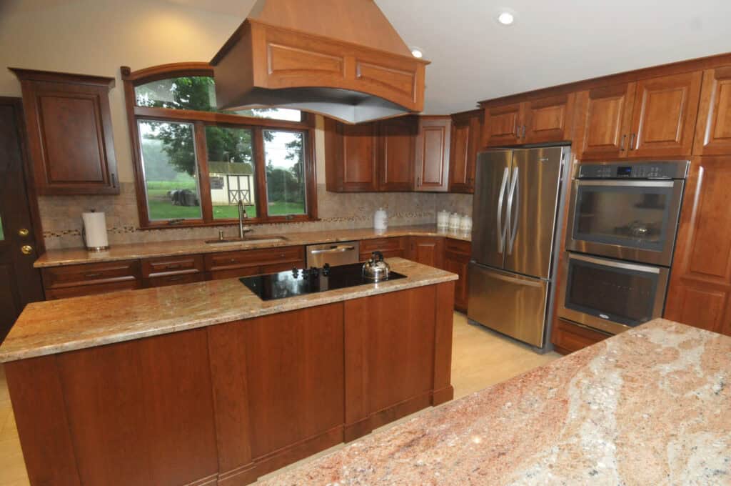 A modern kitchen with wooden cabinets, granite countertops, a kitchen island with a stovetop, stainless steel appliances, double ovens, and a large window overlooking a green yard.