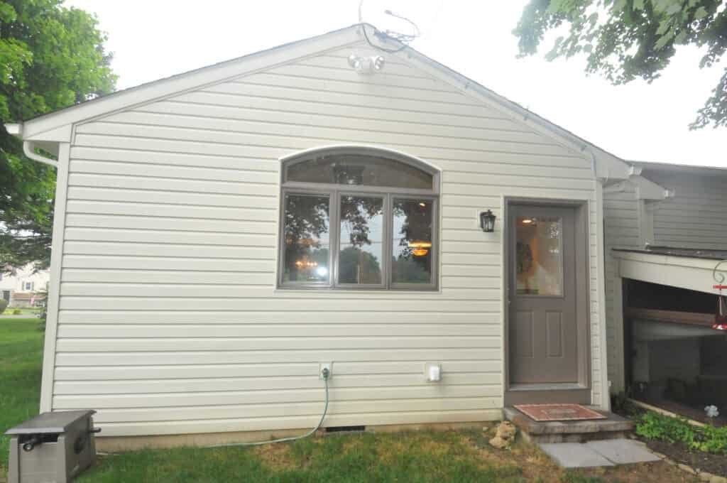The image shows the exterior of a light-colored house with vertical siding, a large arched window, a gray door with a mat, and a green lawn with trees partially visible.