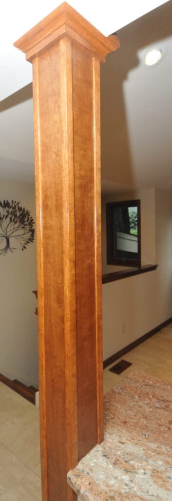 A wooden support column with a smooth, polished finish stands next to a granite countertop in a bright, modern interior with light-colored walls and tiled flooring.