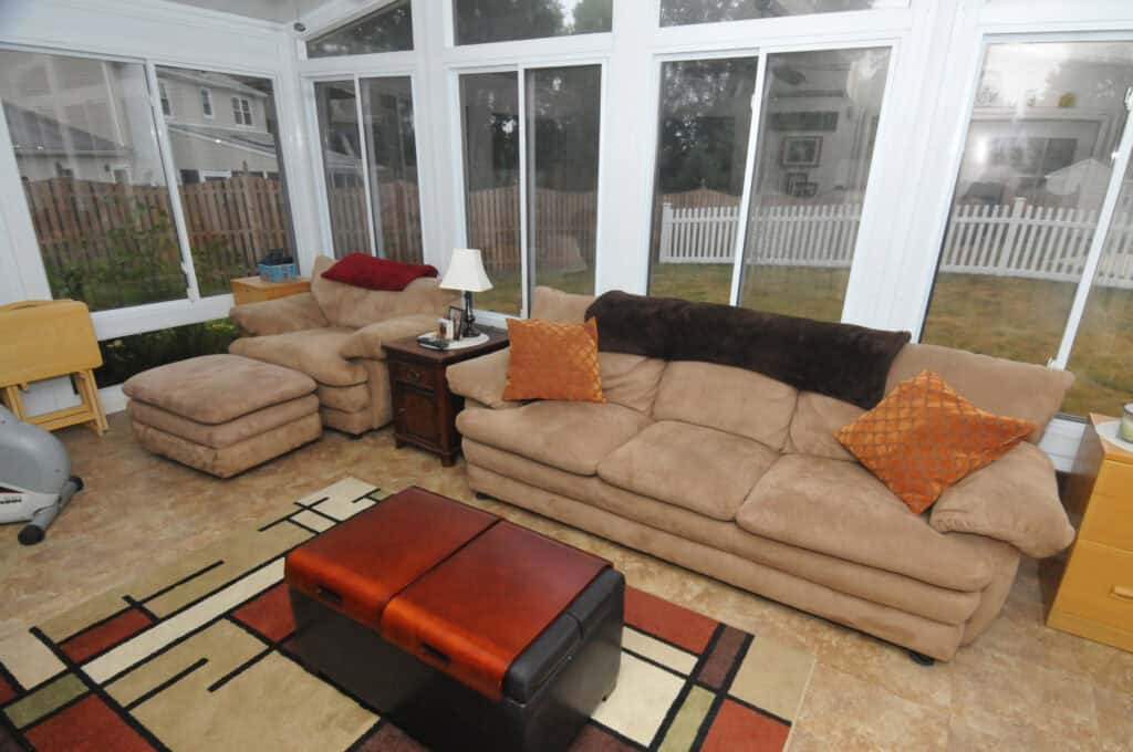 A cozy sunroom with large windows, tan sofas with orange pillows, a matching armchair and ottoman, a small side table with a lamp, a patterned rug, and a two-tone brown ottoman.