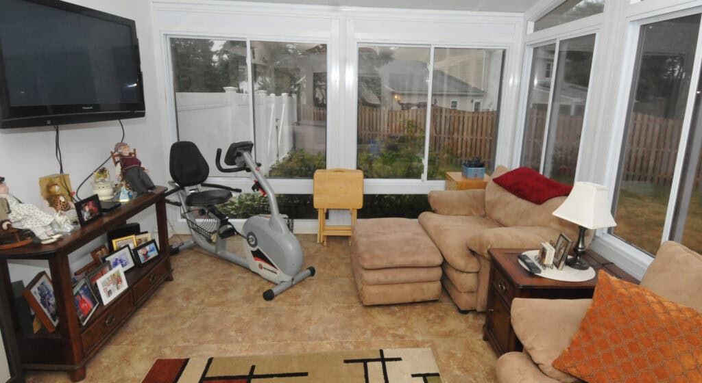 A cozy sunroom with large windows, a stationary exercise bike, a tan armchair with ottoman, a wooden side table with lamp, photos, and a TV mounted on the wall. A beige rug covers part of the floor.
