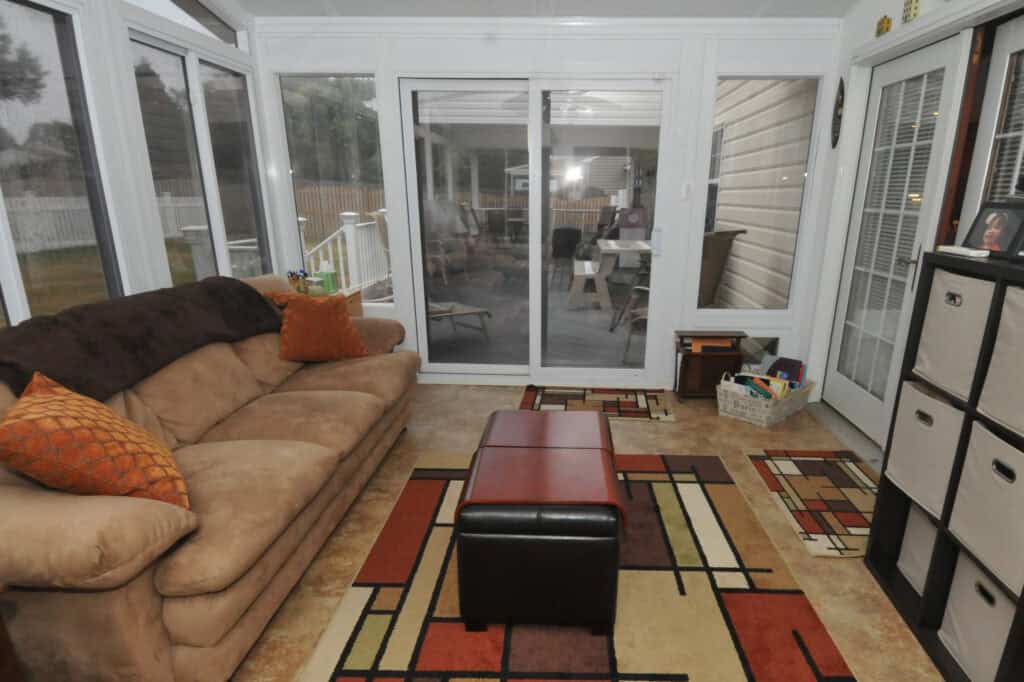 A sunroom with large windows, a beige couch with orange pillows, a brown ottoman, colorful geometric rugs, storage bins, and sliding glass doors leading to an enclosed porch.
