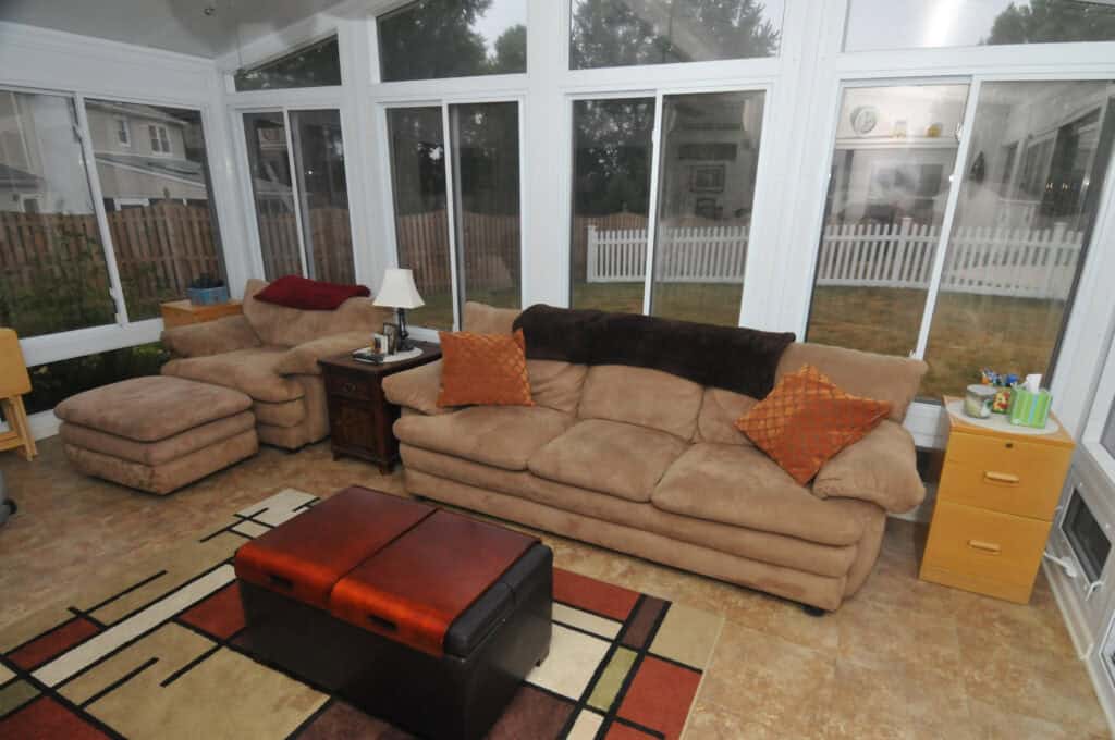 A bright sunroom with large windows features a beige sofa and armchair with orange pillows, a patterned rug, a brown ottoman, and side tables with lamps and decor. A fenced yard is visible outside.