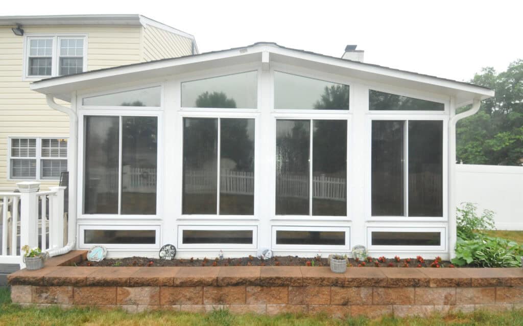 A white sunroom with large windows and a peaked roof extends from a house. In front, a raised brick flower bed holds small plants, flowers, and decorative plates. A white fence and lawn are visible in the background.
