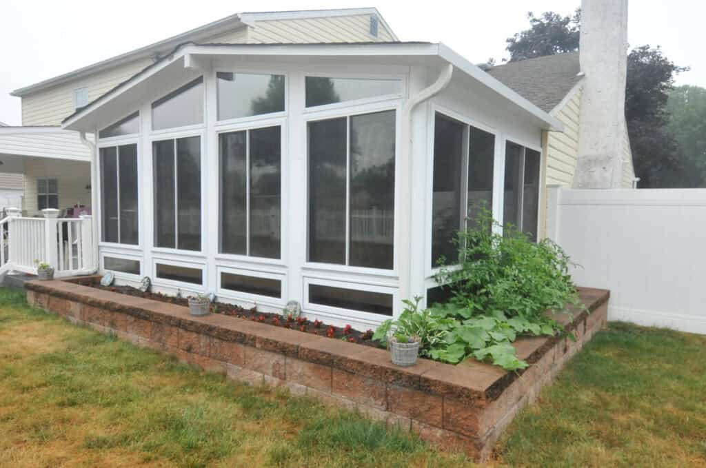 A sunroom with large windows sits on top of a raised brick flower bed, which contains green plants and small potted flowers, next to a beige house with a fenced yard.