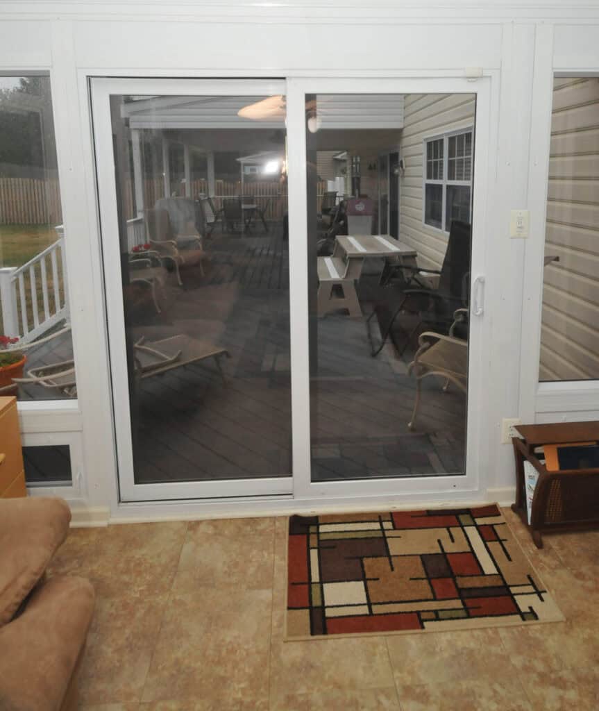 A sliding glass door opens onto a covered porch with patio furniture. Inside, there is tiled flooring, a brown couch, a patterned rug, and a wooden magazine rack. The porch has a wooden floor and beige siding.
