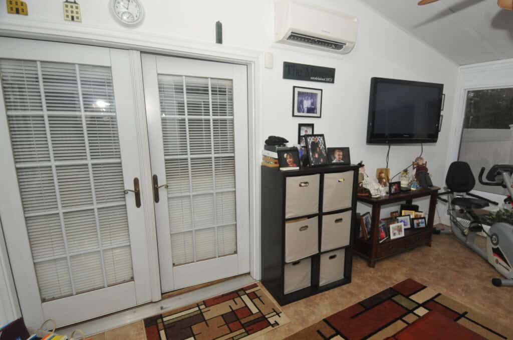 A home office with French doors, a wall-mounted TV, storage cubbies, framed photos, and an exercise bike. A patterned rug is on the floor, and an air conditioner is above the door.