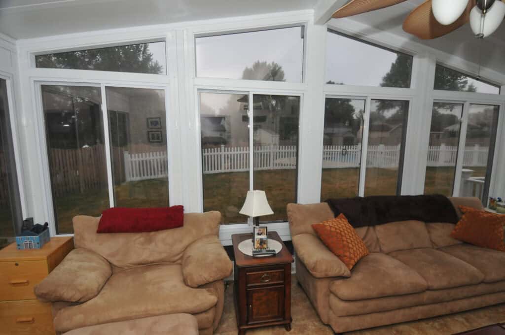 A sunroom with large windows shows an outside view of a fenced yard. Inside, there are two beige couches with colorful pillows, a wooden side table holding a lamp and picture frame, and a ceiling fan overhead.