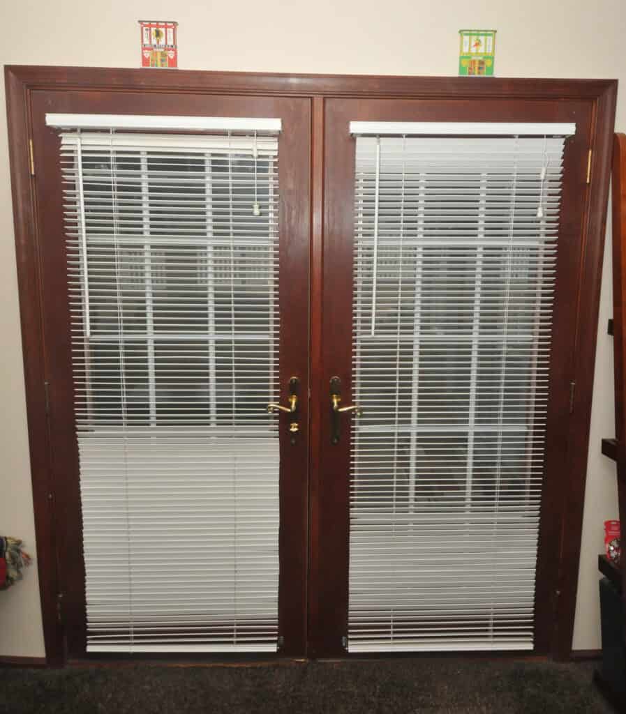 A set of double glass doors with dark wooden frames, each door fitted with white horizontal blinds. Two small decorative lanterns sit above the doors, one red and one green.