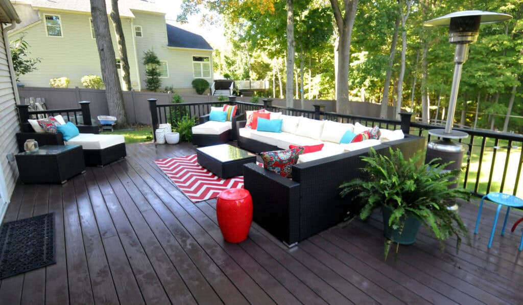 Spacious outdoor deck with wicker sofas, white cushions, colorful pillows, a red and white chevron rug, potted plants, a red ceramic stool, and a patio heater, surrounded by trees and a fenced yard.