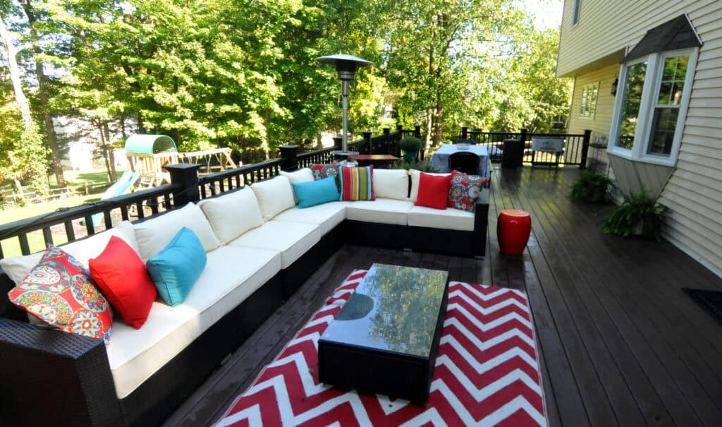 A spacious wooden deck features a wicker sectional sofa with white cushions and colorful pillows, a glass coffee table, and a red-and-white chevron rug. Trees and a playground are visible in the background.