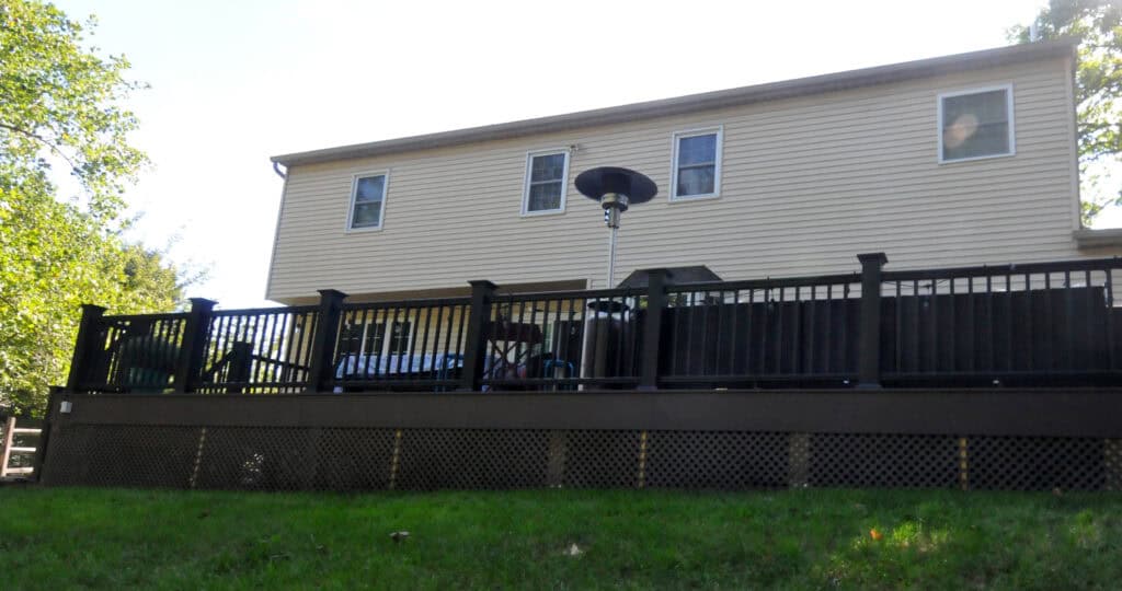 A two-story beige house with white trim and four windows, featuring a raised dark-colored deck with a black railing and a patio heater, overlooking a grassy yard.