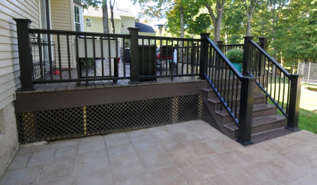 A backyard deck with black railings and stairs leads down to a tiled patio. The deck features lattice skirting along the base, and the area is surrounded by trees and greenery.