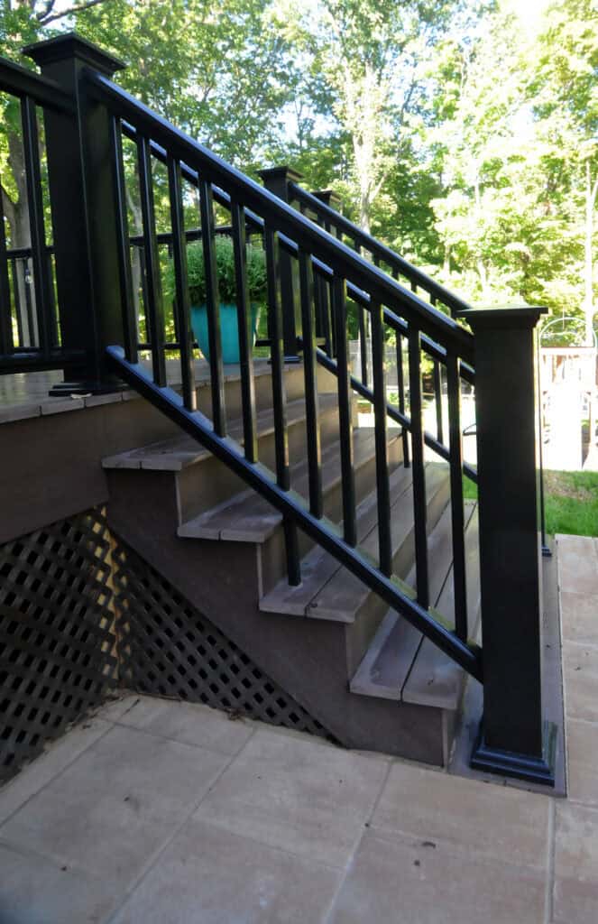 A wooden outdoor staircase with dark railings leads from a tiled patio up to a deck. Sunlight filters through trees in the background, and a potted plant sits on the deck above the stairs.