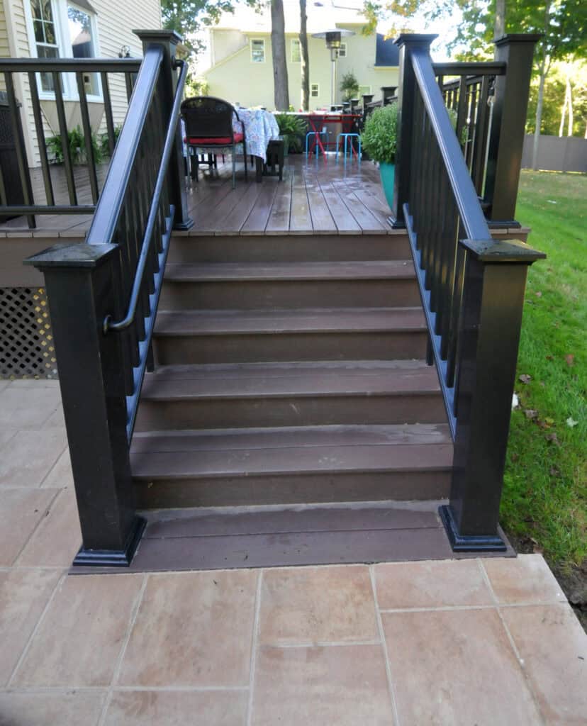 A set of brown wooden stairs with black railings leads from a tiled patio up to a raised deck, where outdoor furniture and plants are visible in a backyard setting.
