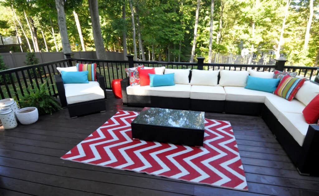 An outdoor patio with a black wicker sectional sofa, white cushions, and colorful throw pillows. A red and white chevron rug and a black coffee table are on dark wood decking, surrounded by trees and greenery.
