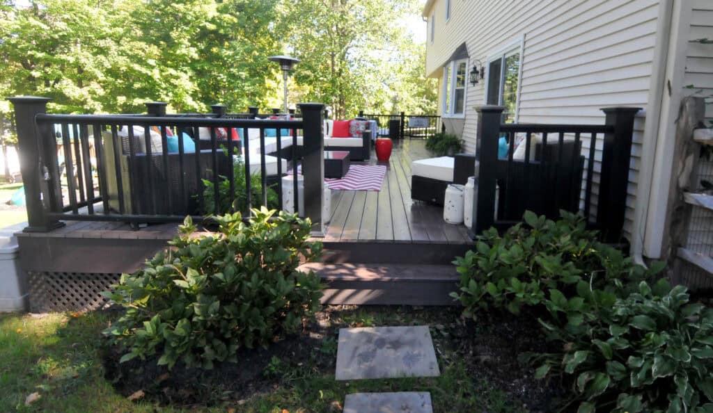 A backyard deck with black railings and outdoor furniture, including a sofa and a red and white striped blanket, surrounded by green plants and trees on a sunny day.
