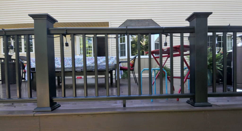 A black metal railing encloses a deck with outdoor furniture, including a patterned cushioned bench, a red table, and colorful chairs, in front of a beige house with windows.