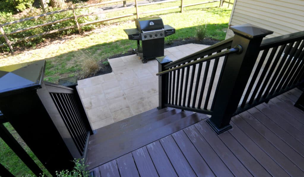 View from a wooden deck with black railings, looking down stairs to a paved patio with a barbecue grill and grass yard bordered by a wooden fence.