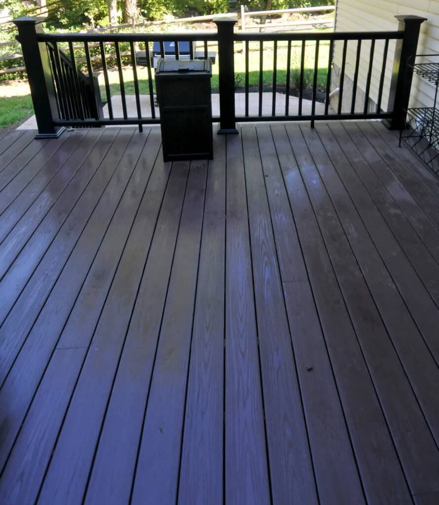 A clean, empty wooden deck with black railings, a square trash bin, and a view of a green lawn and trees in the background. Sunlight casts shadows across the deck floor.