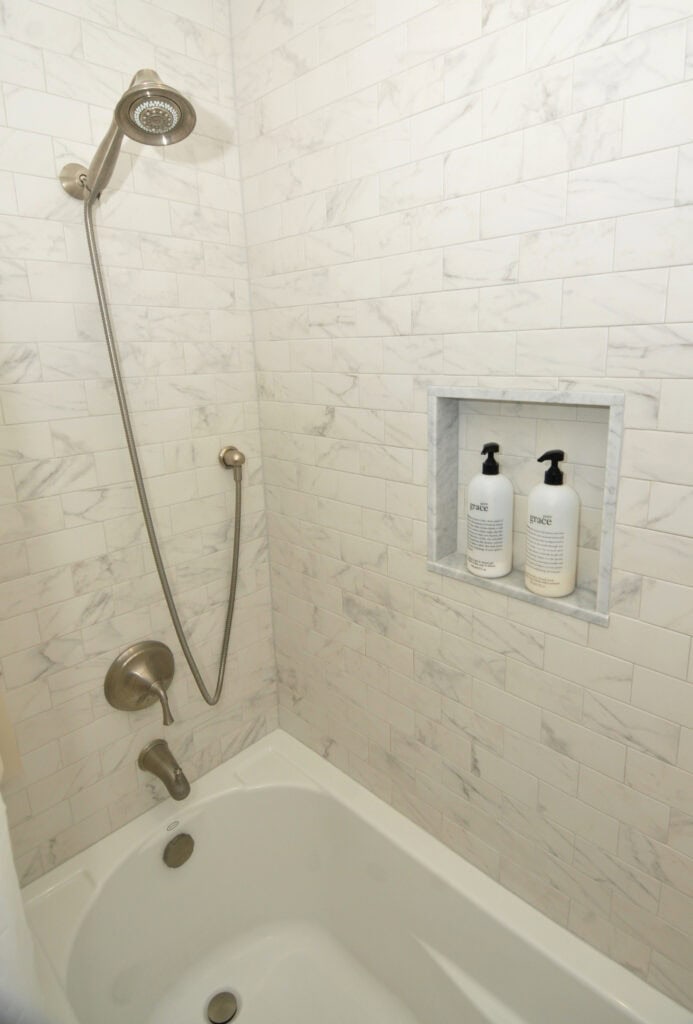 A white marble-tiled shower with a bathtub, a silver showerhead, faucet, and a built-in niche holding two bottles of soap or shampoo.