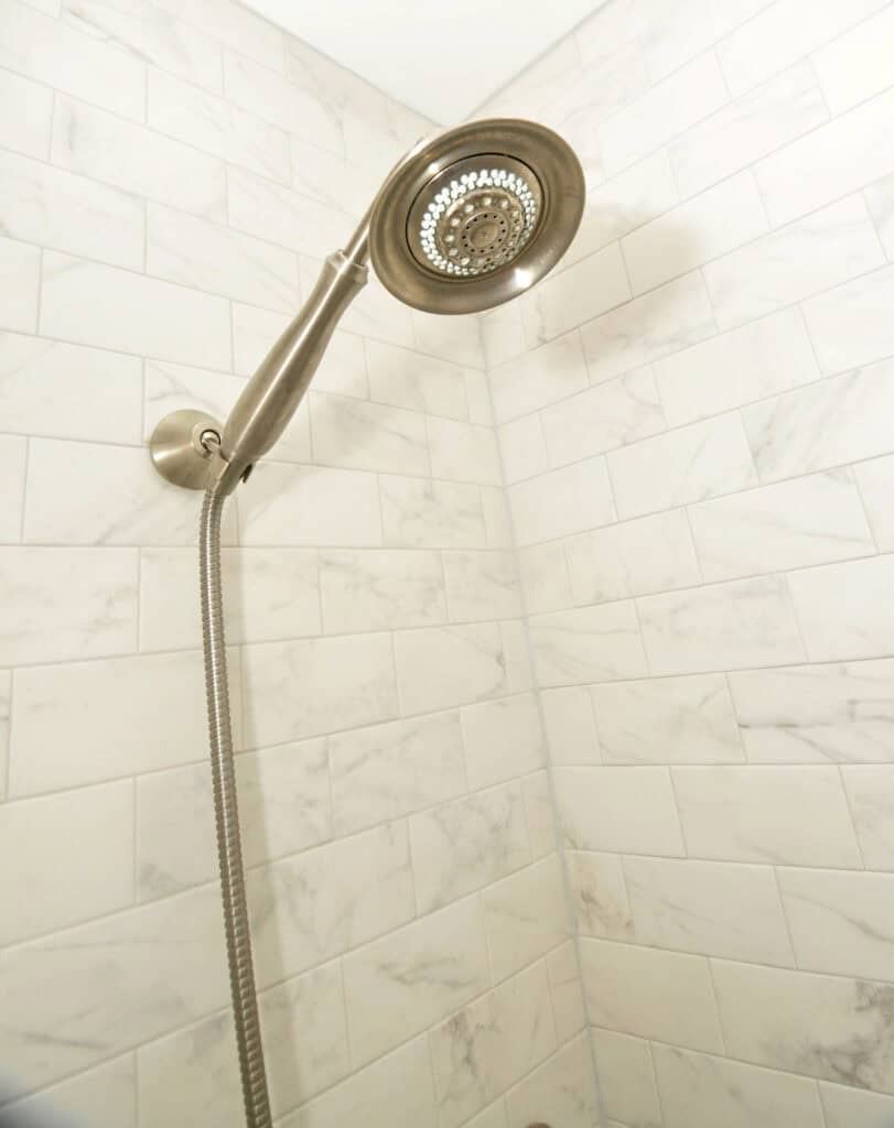 A stainless steel handheld showerhead is mounted on a tiled shower wall with white, marble-patterned tiles arranged in a subway style.