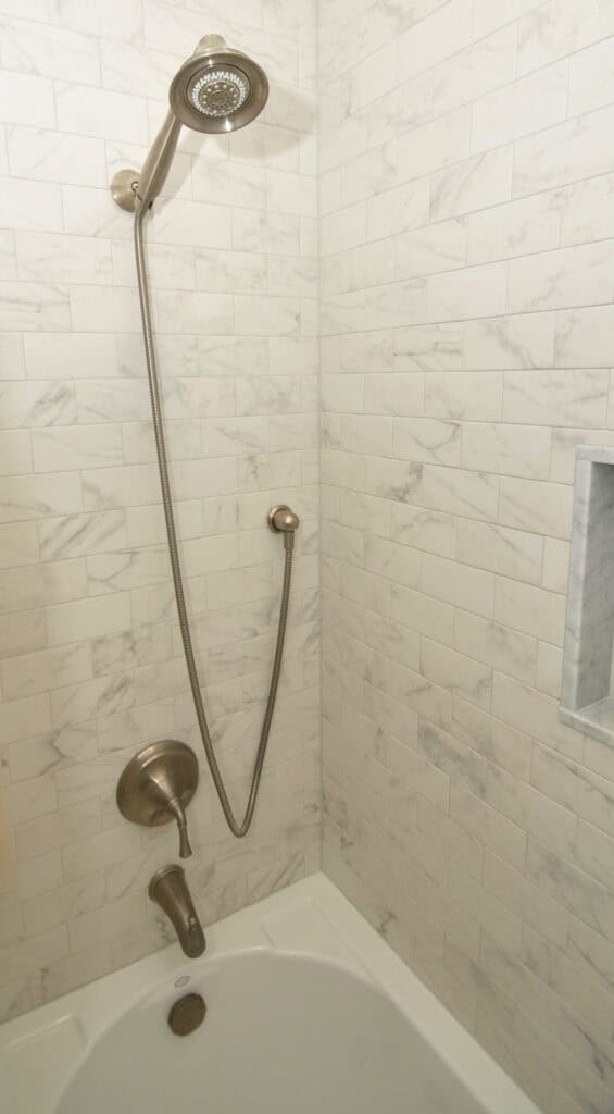 A tiled shower with a bathtub, featuring a handheld showerhead on a long hose, a wall-mounted faucet, a control knob, and a recessed shelf built into the tiled wall.