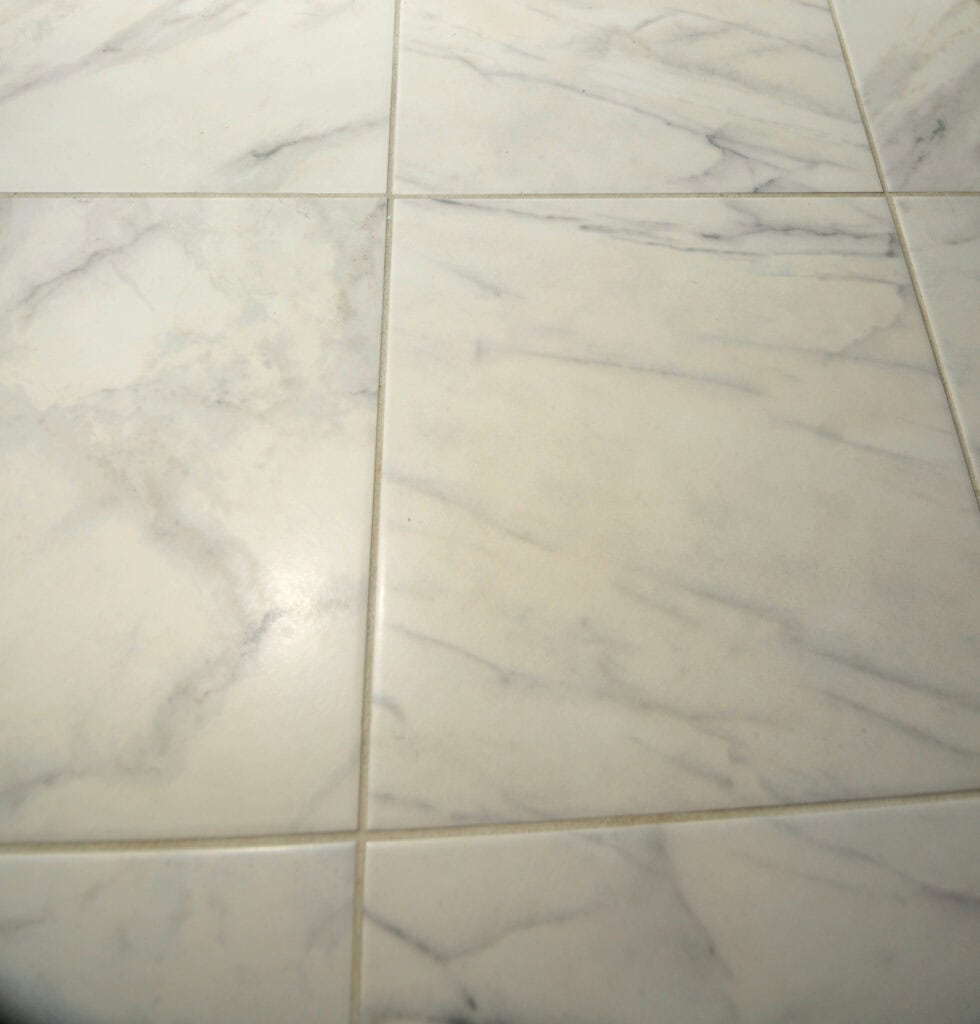 Close-up of light-colored marble tiles with gray veining and grout lines, showing a smooth, polished surface and subtle natural patterns.