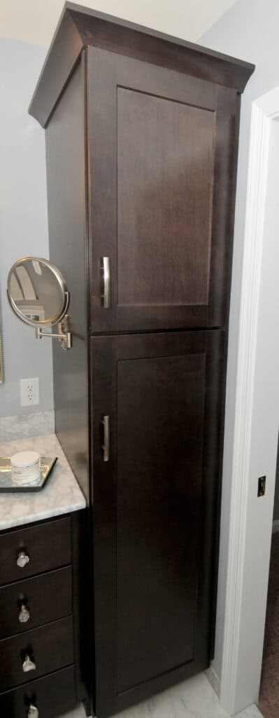 Tall, dark wooden bathroom cabinet with two doors and silver handles, next to a marble countertop with a tray and a wall-mounted round mirror.