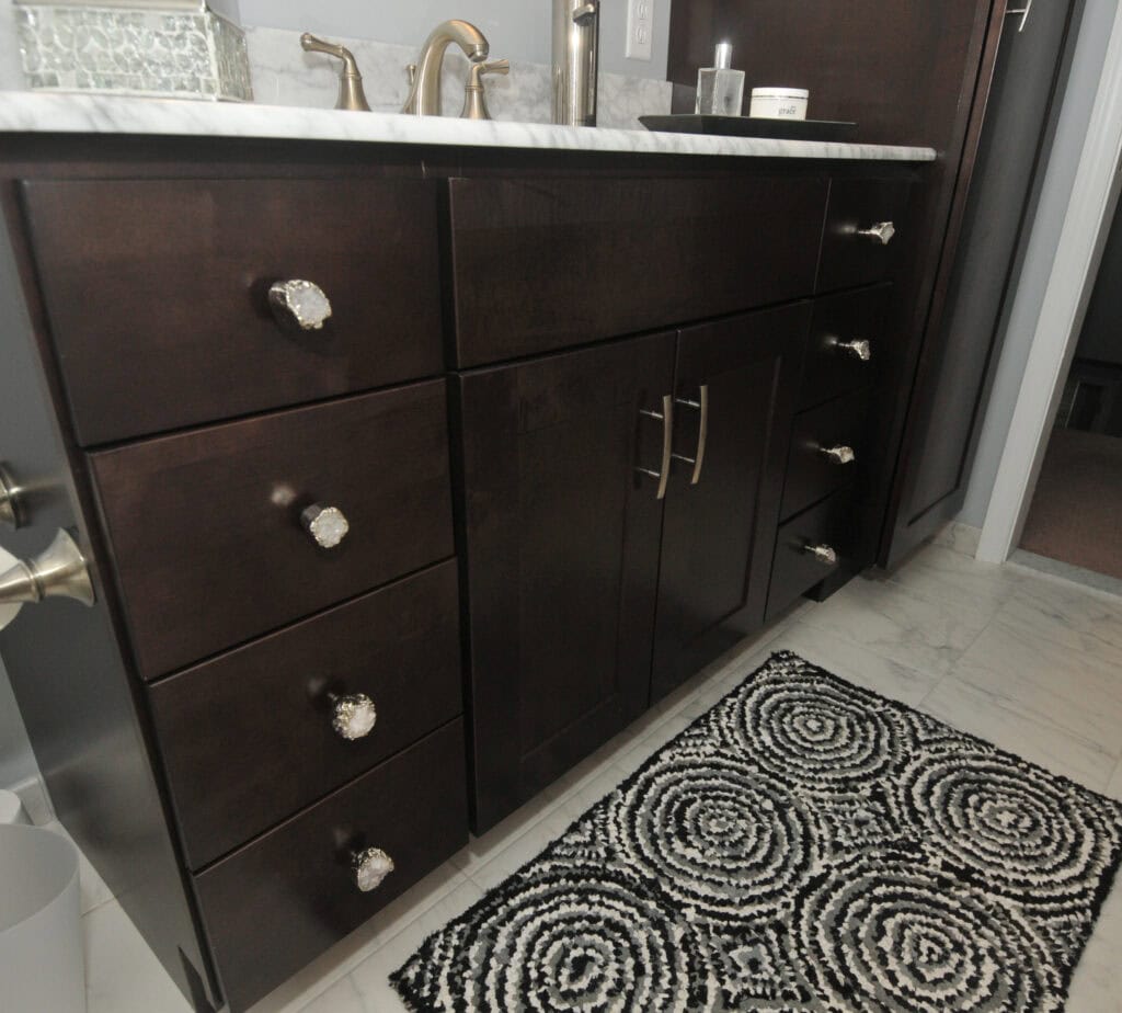 Dark wood bathroom vanity with silver hardware, marble countertop, silver faucet, and decorative items. A black and white textured rug with a circular pattern lies on the tiled floor in front.