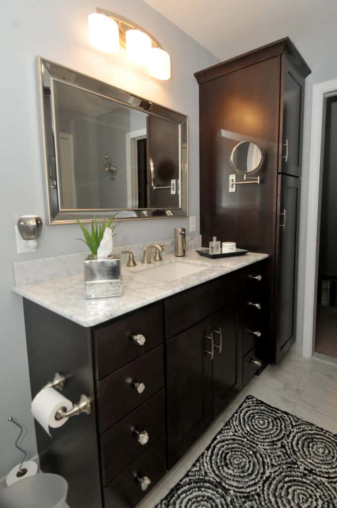 Modern bathroom with a dark wood vanity, marble countertop, large mirror, and wall-mounted lights. Decorative items and a plant are on the counter. A tall cabinet and patterned rug are also visible.