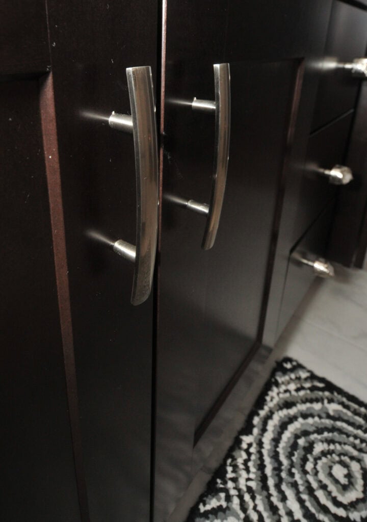 Close-up of dark brown cabinet doors with silver handles, next to drawers with matching knobs. A black and white patterned rug is visible on the floor in the lower right corner.