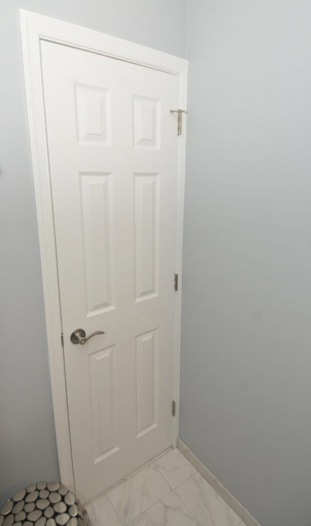 A white, paneled interior door with a silver handle and top security latch, set in a light gray wall with white trim and marble-tiled floor.